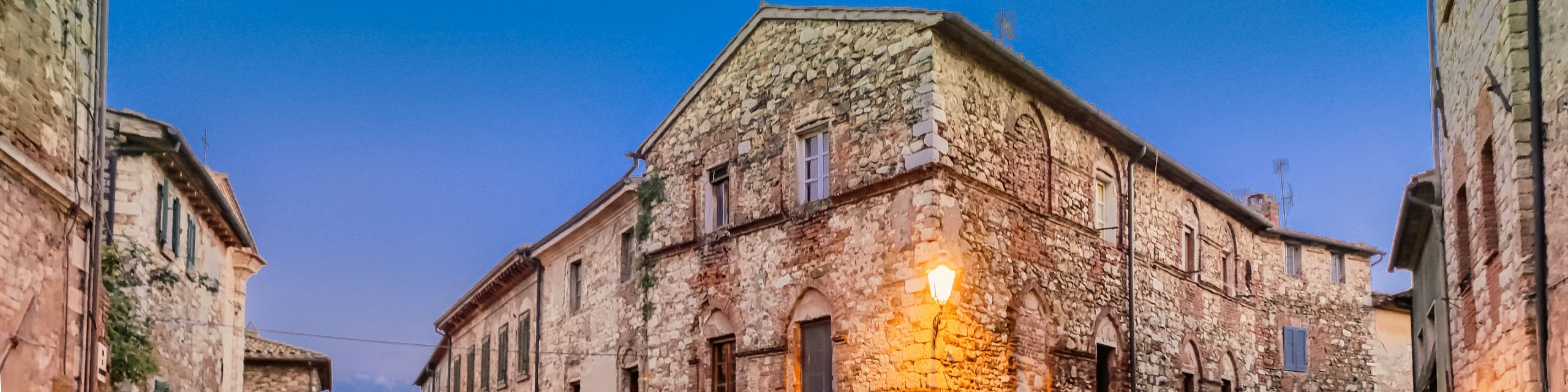 Looking up at a series of medieval stone buildings with one brightly lit street lamp and a dark blue twilight sky